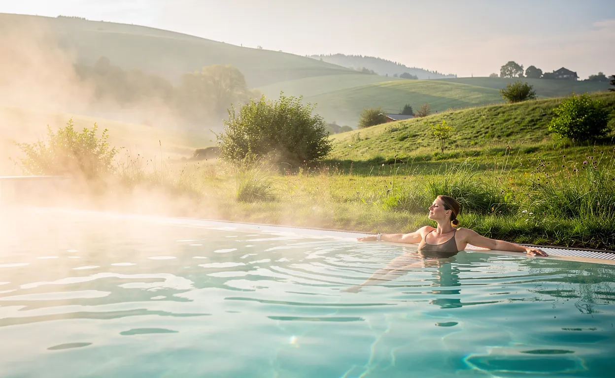 Une personne se détend au bord d'un bassin thermal extérieur entouré de collines verdoyantes du Jura, avec de la vapeur s'élevant dans une atmosphère paisible au petit matin.