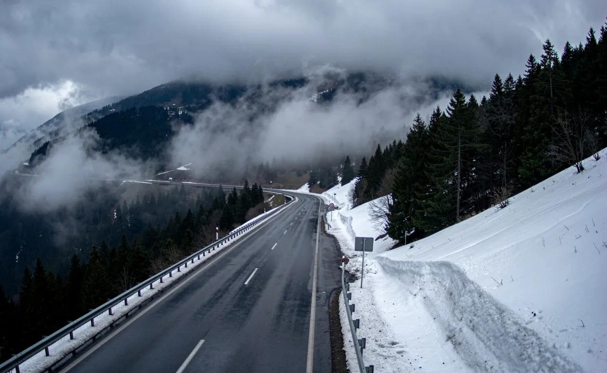 Col de la Faucille sous un ciel nuageux avec route sinueuse, pentes enneigées et brume parmi les pins.