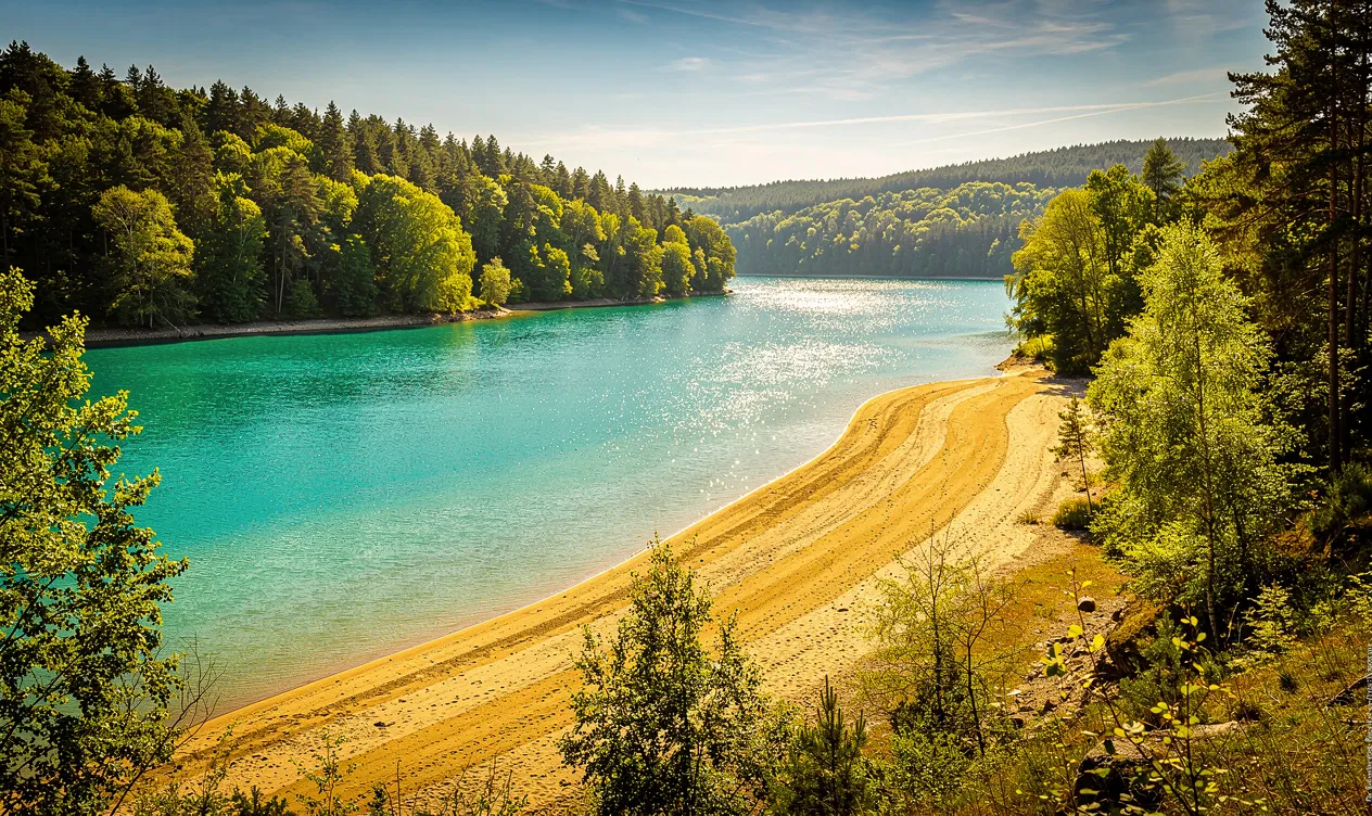 Lac de Vouglans aux eaux turquoise bordé de plages dorées et de forêts verdoyantes sous un ciel d’été lumineux.
